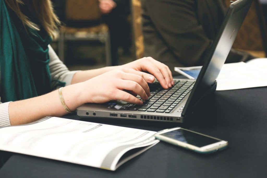 Adult woman typing on a laptop at a desk with a smartphone and documents.